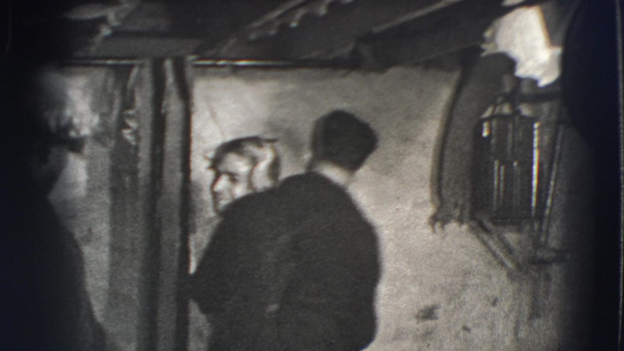 Adult men in a dim basement, one turned away as another leans near a rough wall.