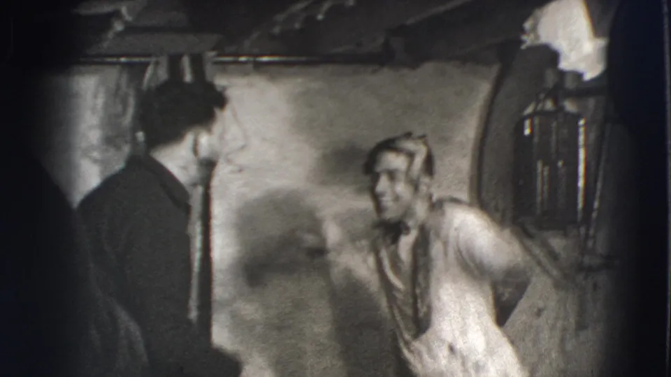 Joliet, Illinois 1941 frame with a shirtless adult man smiling and leaning forward in a dim basement beside a man in dark clothing.