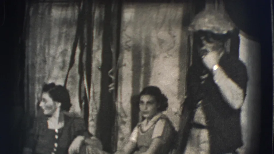 Women seated in a dim Joliet interior watch a male performer holding a light pipe or prop near draped curtains.