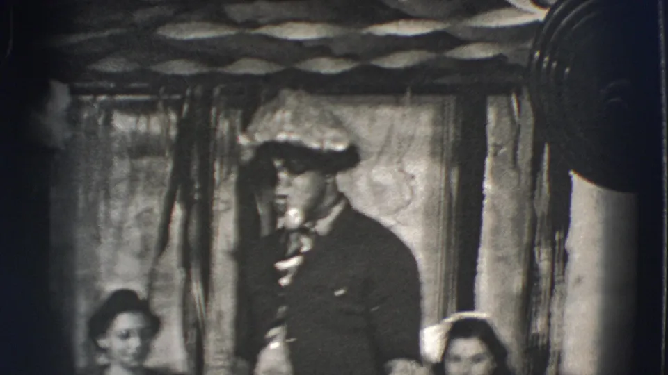A 1941 Joliet stage performer in a brimmed hat holds a white pipe, with seated women partly visible in the foreground.