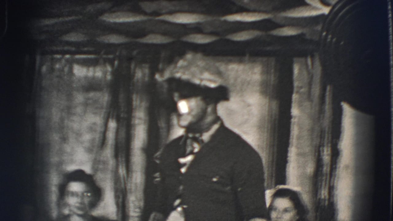 Man in suit and hat stands indoors while seated people watch from shadow.