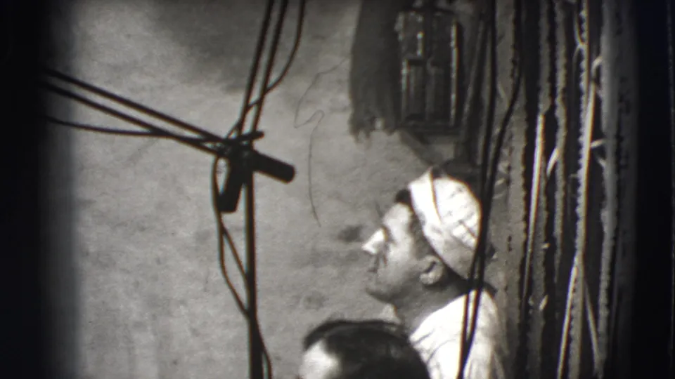 1941 Joliet frame showing a side-profile man in a white cap beside hanging wires, rough wall texture, and a wall-mounted fixture.