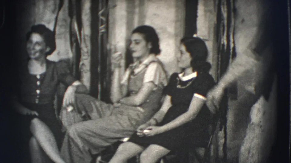 The home-movie frame shows three young women seated in a Joliet backyard or courtyard as someone moves through the foreground.
