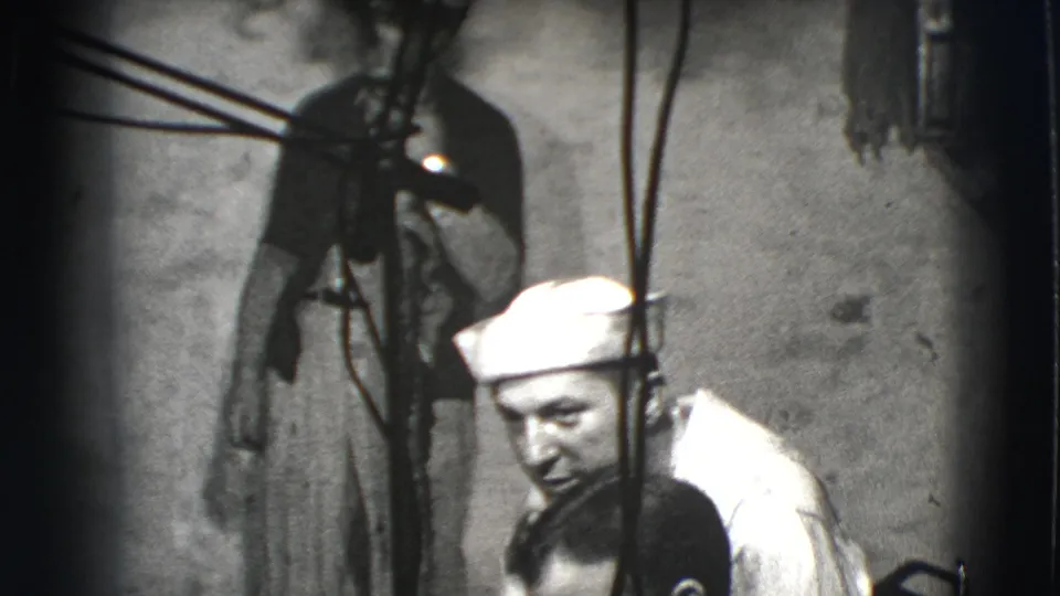 1941 Joliet home-movie frame showing a man in a white sailor cap leaning near another man amid hanging cords in a dark interior.