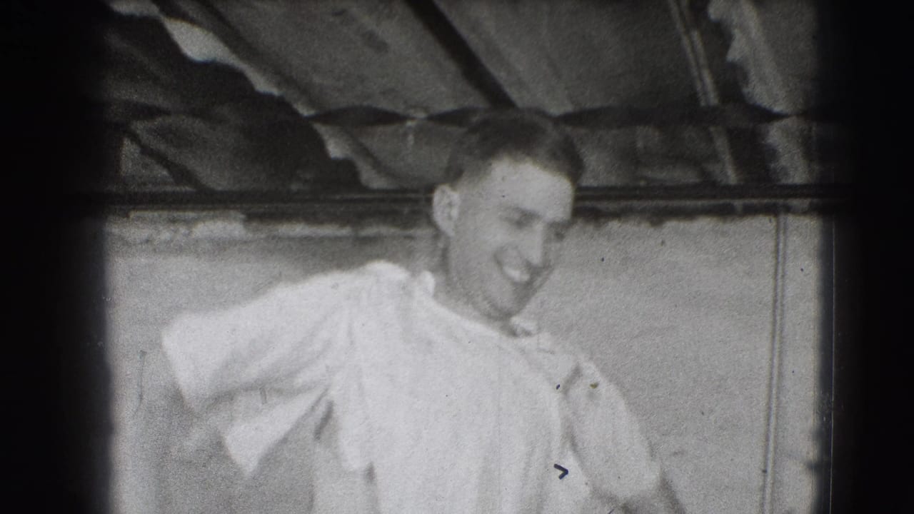 Smiling man in a light shirt moves in a dim room with exposed beams overhead.