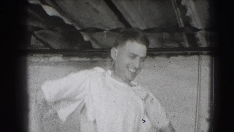 In 1941 Joliet home-movie footage, a smiling adult man in a light shirt moves near the camera inside a plain, beam-lined interior.