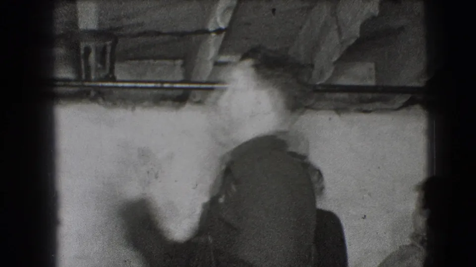 A blurred adult in dark clothing turns or gestures near a plain wall, with exposed ceiling structure visible above.