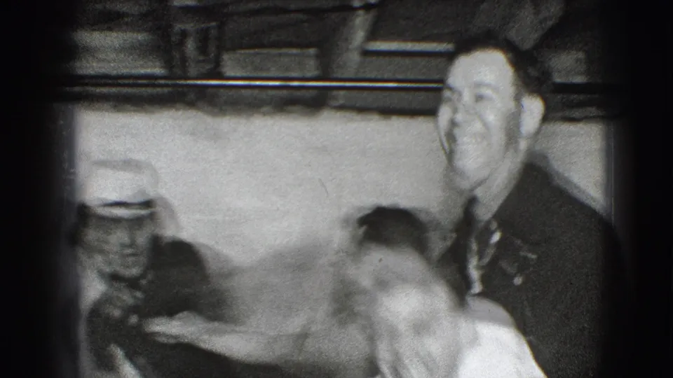 A smiling adult man in a dark jacket stands near blurred companions, including a figure in a light hat, in the 1941 Joliet indoor scene.