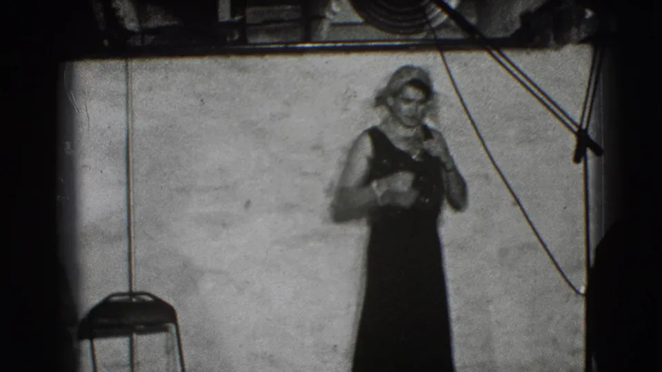A costumed adult in a dark sleeveless dress stands against a plain wall in the Joliet rehearsal space, with stage equipment visible above.
