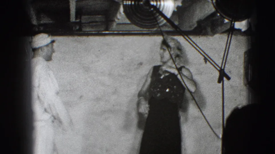 A light-clothed adult observes or speaks with the costumed performer near visible cables and a plain wall in the Joliet rehearsal setting.