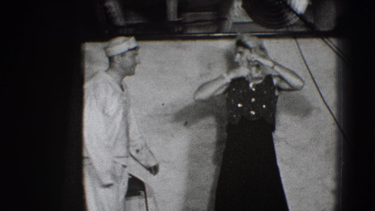 Man in a white uniform watches a woman in a dark dress handle a hat on stage.