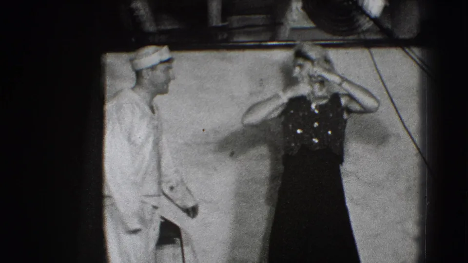 In 1941 Joliet, Illinois footage, a woman in a dark stage dress holds a hat while a man in a white uniform stands nearby.