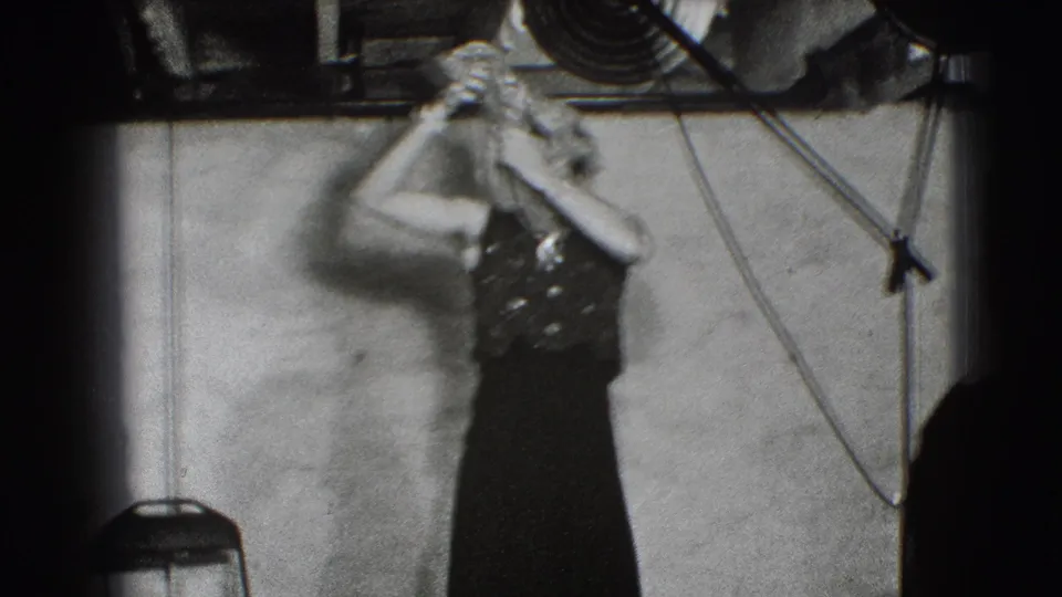 A woman in a dark sequined dress raises a hat during a stage magic routine in the Joliet, Illinois home-movie clip.