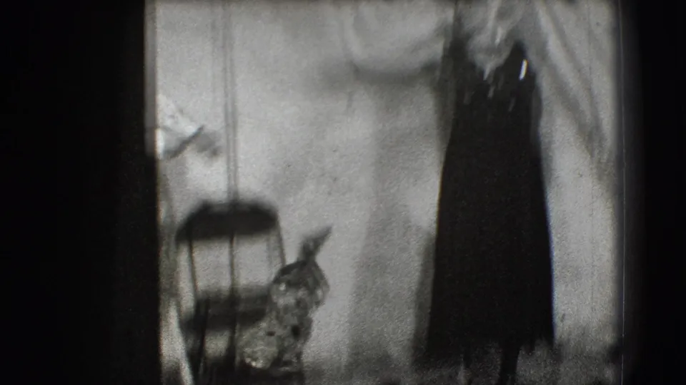A soft, close 8mm frame shows the performer moving with the hat prop, with a chair and stage wall partly visible.