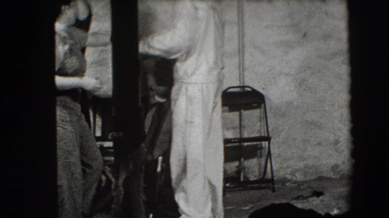 Adults stand in a dim room beside a folding chair and rough wall.