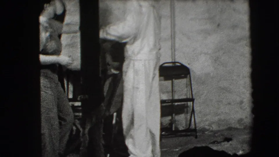 In a Joliet, Illinois 1941 home-movie frame, adults stand partly cropped in a dim interior beside a folding chair and plain wall.