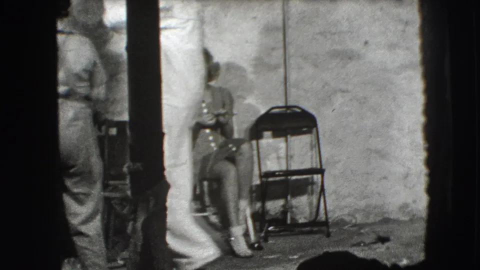 A white-uniformed adult stands in a plain Joliet interior while a woman sits nearby beside a folding chair.