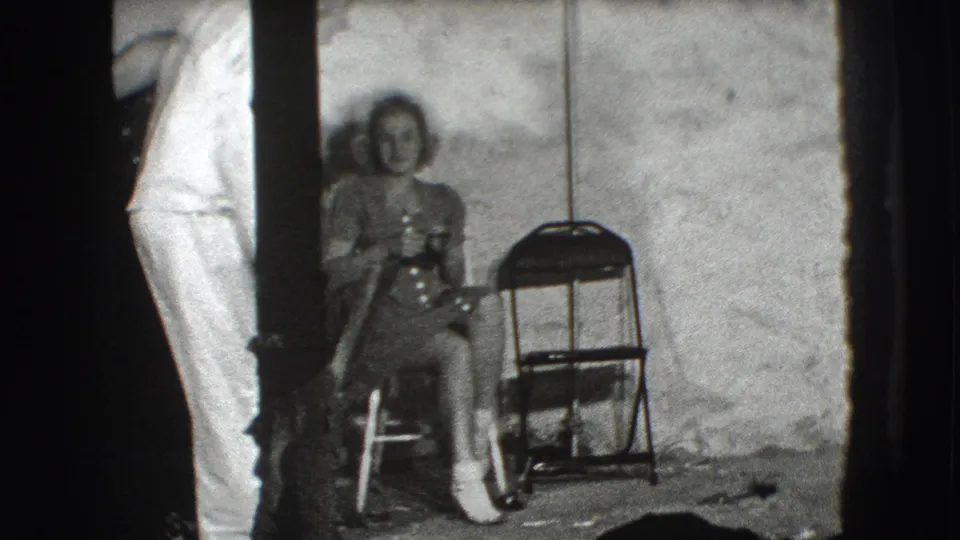 A seated woman in a Joliet 1941 interior faces the camera as standing adults pass near a folding chair and rough wall.