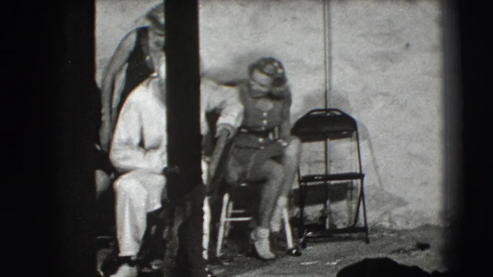 In the Joliet interior, a woman in overalls sits beside a folding chair while a white-uniformed adult leans or sits close to her.