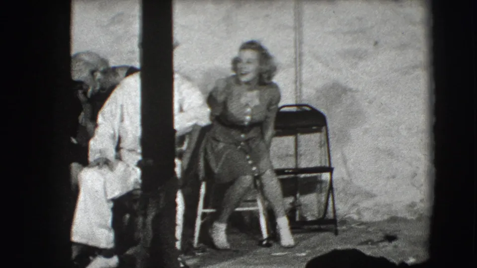 In 1941 Joliet, Illinois footage, a young woman sits on a folding chair as sailors in white uniforms stand close by in a studio setting.