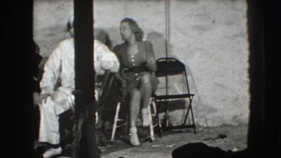 In the Joliet studio scene, the woman remains seated and smiling while sailors in white uniforms move near her within the dark film frame.