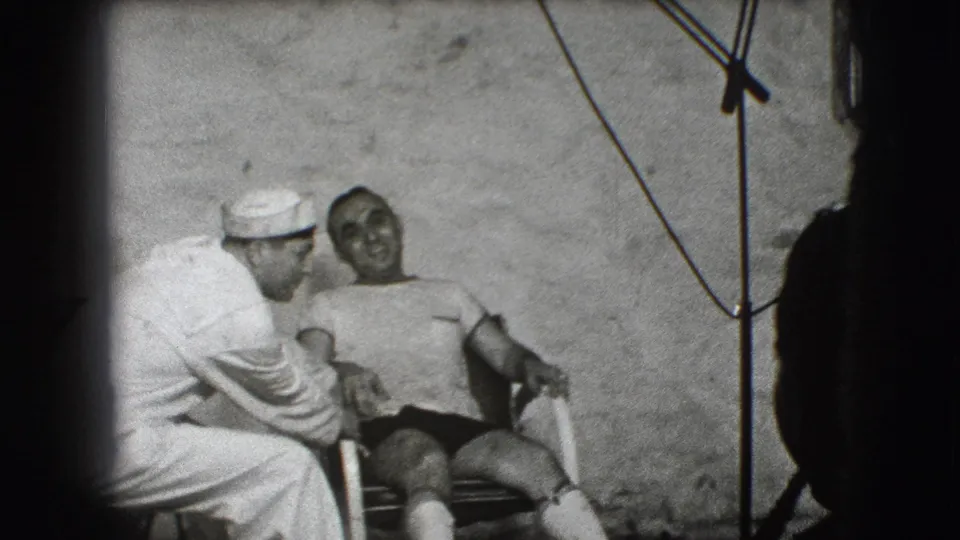 A sailor in white bends toward a seated man in shorts, with the woman partly visible behind them against the Joliet studio backdrop.