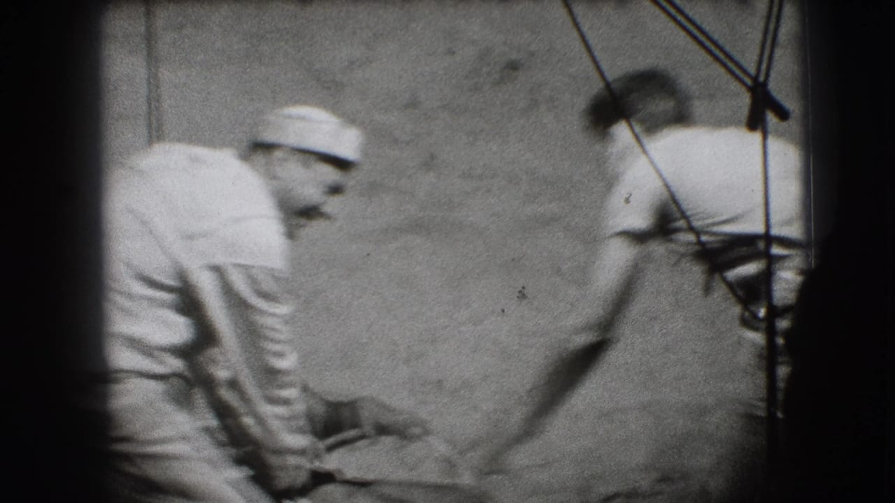 Man in a white cap leans toward another adult beside a wheelchair in black-and-white home-movie footage.