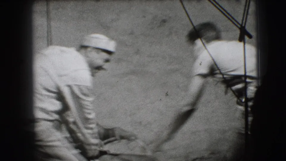 In 1941 Joliet home-movie footage, a man in a white cap leans toward another adult near a wheelchair against a plain wall.
