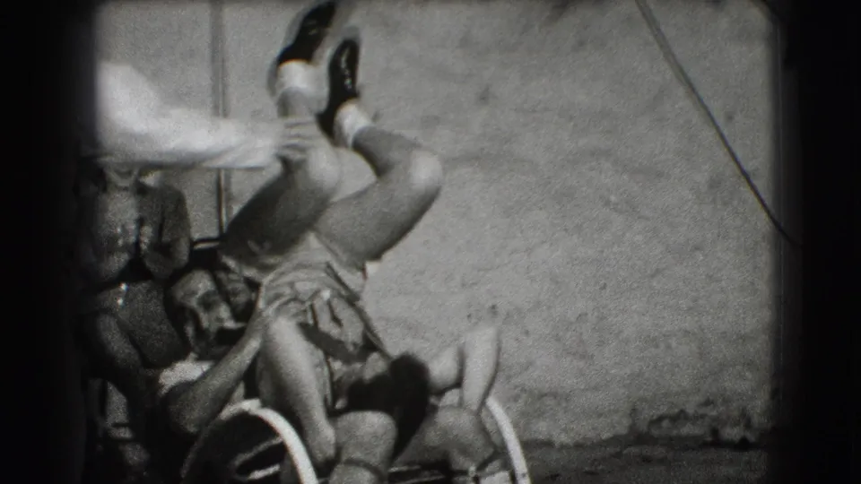 Adults in the Joliet 1941 home movie crowd around a wheelchair as one man's legs lift upward during joking activity.