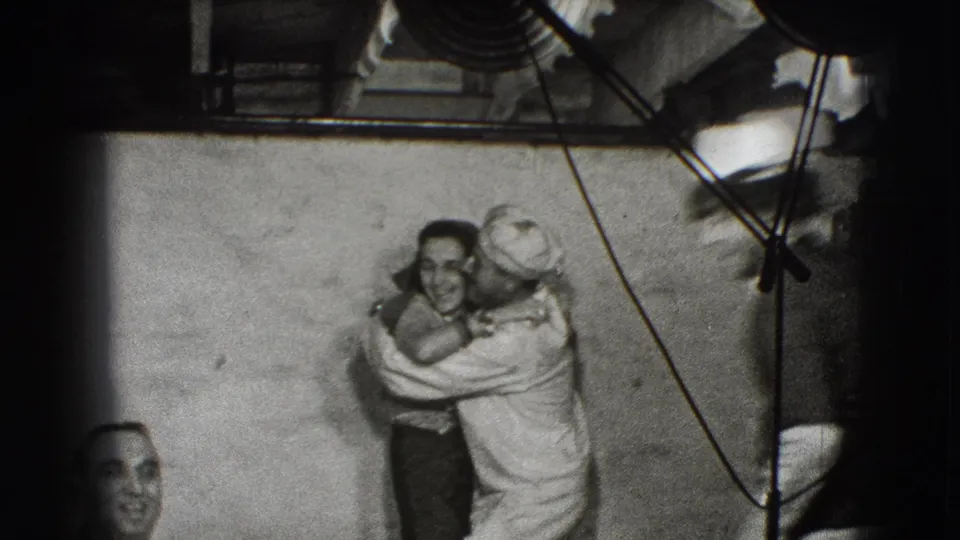 The sailor hugs a woman beside a plain wall or bulkhead, with ropes or cables and metal fittings suggesting a ship or dock setting.