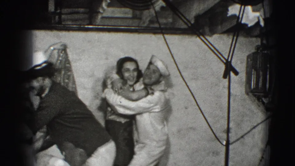 A 1940s home-movie frame shows the sailor embracing the woman while a nearby adult bends forward and rigging-like lines cross the scene.