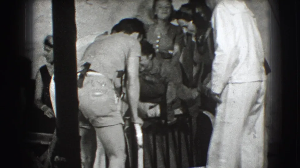 In Joliet, Illinois home-movie footage from 1941, several people stand around as a man bends toward a large metal-framed object indoors.