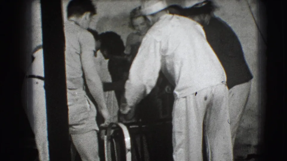 A white-clad man leans in with others to maneuver a metal-framed object in a crowded Joliet interior, recorded on 1941 8mm film.