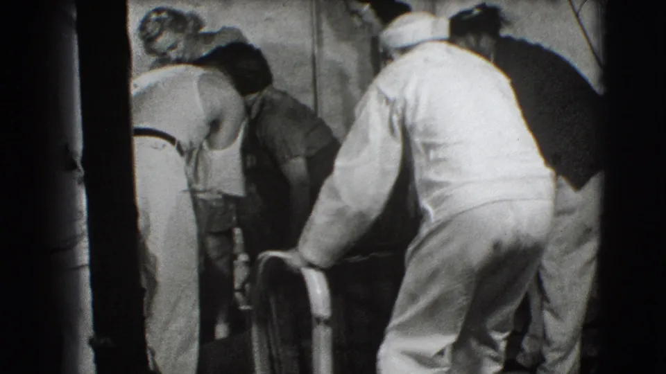 Several adults in 1940s clothing lean over a large metal frame while others watch in the crowded Joliet indoor scene.