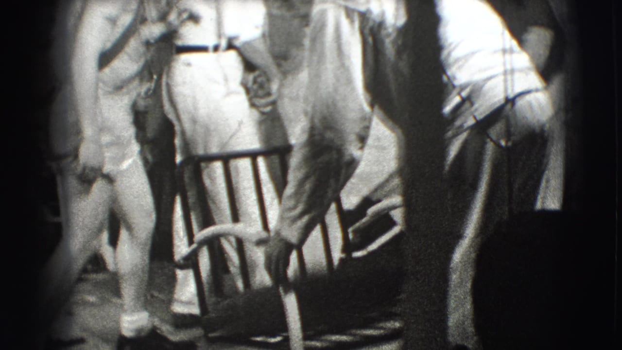 Adults standing around a metal chair in a dim room.