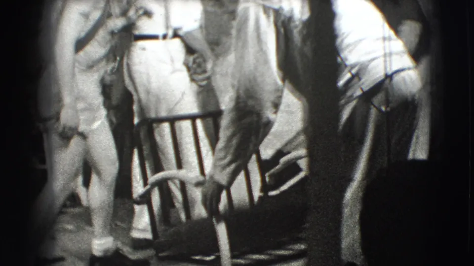 In 1941 Joliet, Illinois home-movie footage, several adults stand close together near a metal chair in a dim interior, with only partial bodies visible.