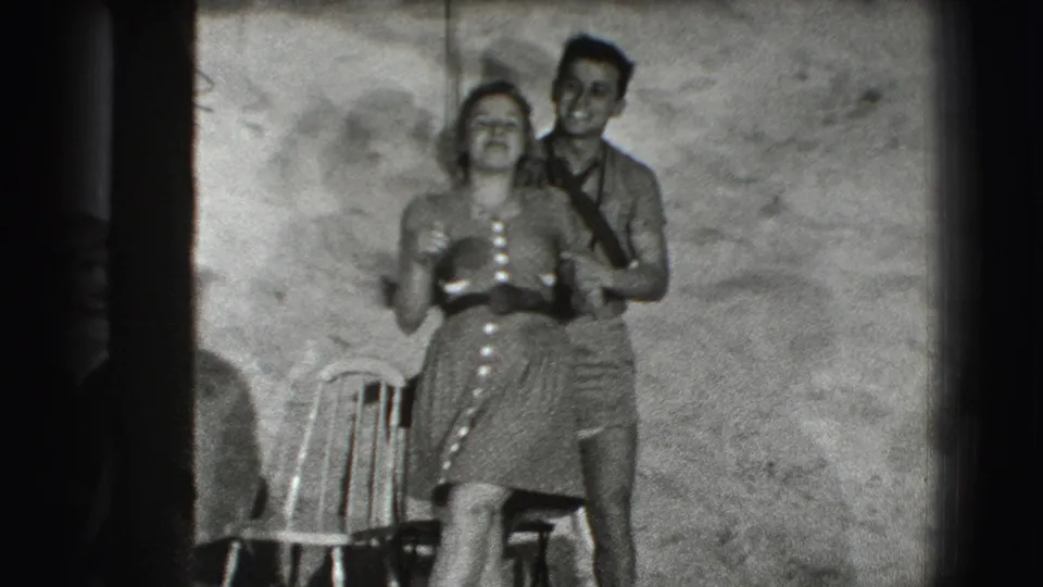 In this 1941 Joliet home-movie frame, a smiling man stands behind a woman in a button-front dress beside a simple chair.