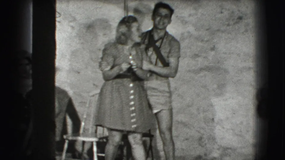 In the Joliet 1941 home-movie frame, the man holds the woman from behind as she turns toward him beside a chair.