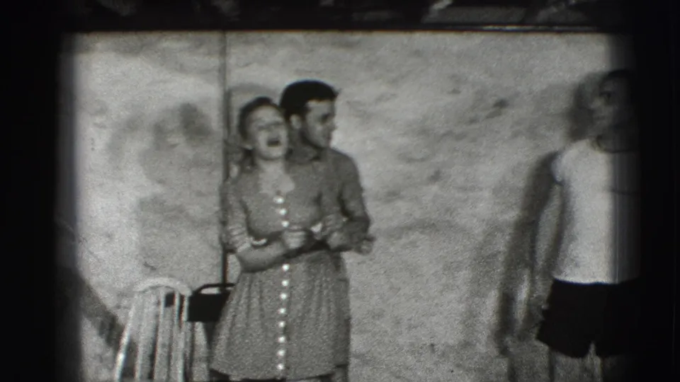 In this 1941 Joliet home-movie frame, a woman in a patterned dress laughs beside a man while another adult stands at the room's edge.