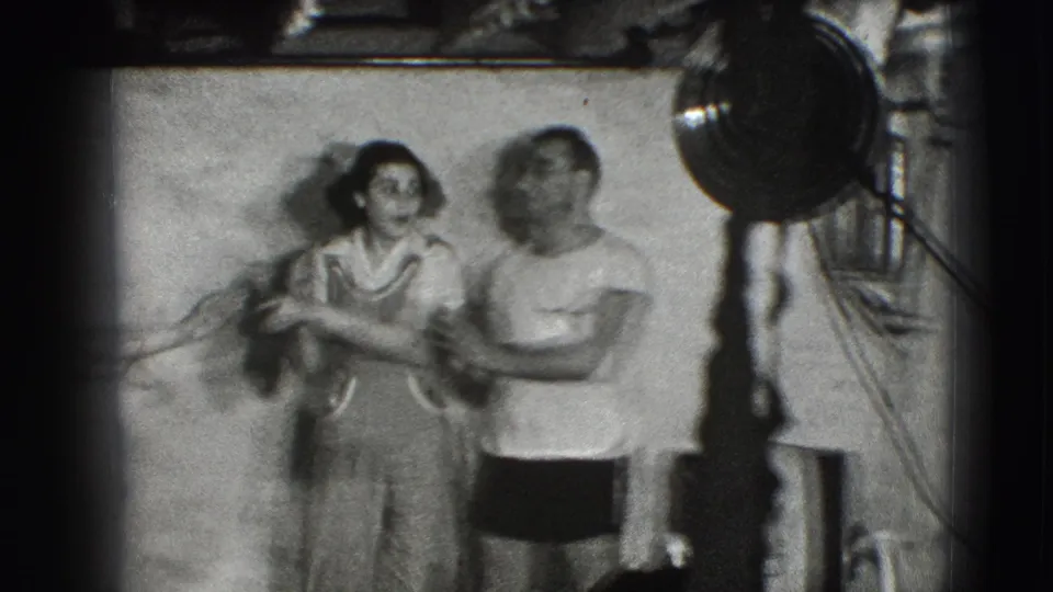 A woman in a dress and a man in a light shirt interact playfully indoors in the supplied 1941 Joliet home-movie footage.