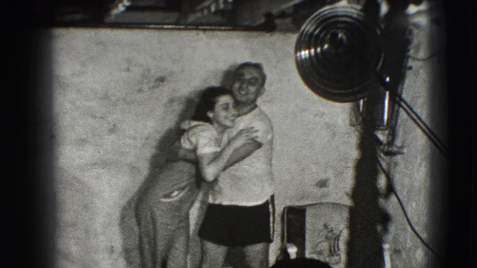 The woman embraces or leans into the man in a casual 1941 Joliet interior, with a large fan and simple furnishings nearby.