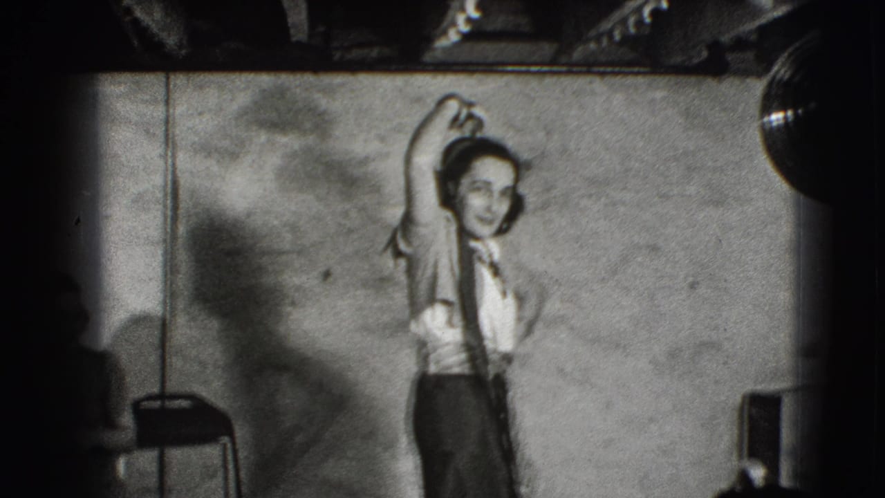 Young woman in a light blouse and dark skirt raises one arm while standing near a chair.