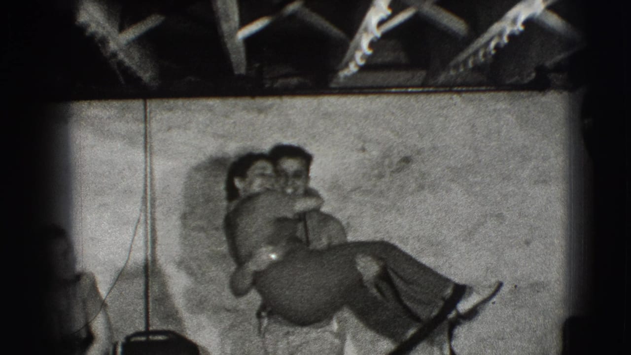 Two young adults embrace closely near a plain wall beneath exposed ceiling beams.