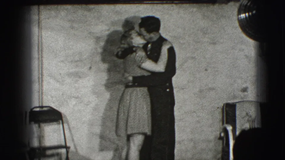 The couple dances face to face in the listed Joliet, Illinois home movie, with a folding chair and plain wall visible nearby.