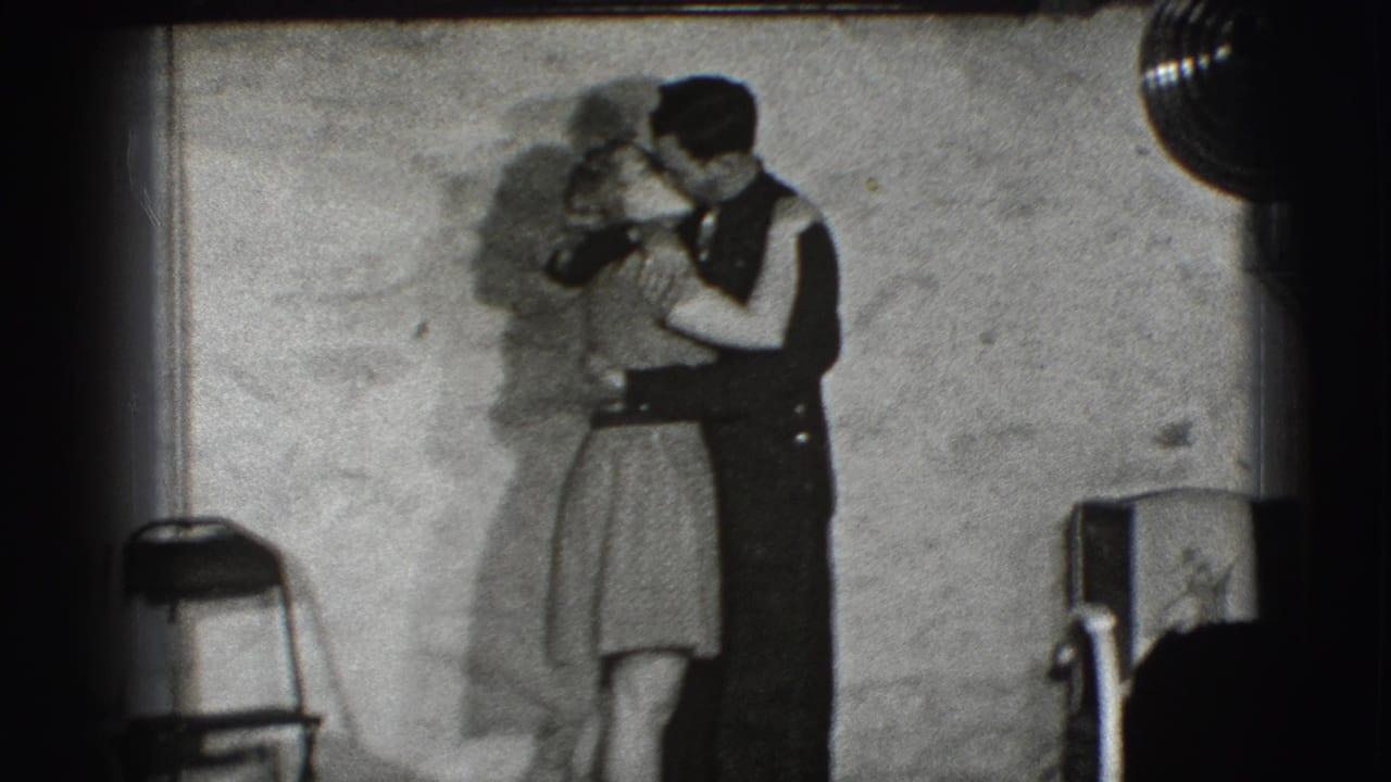 Young couple embraces and kisses beside a plain wall in a dim interior.