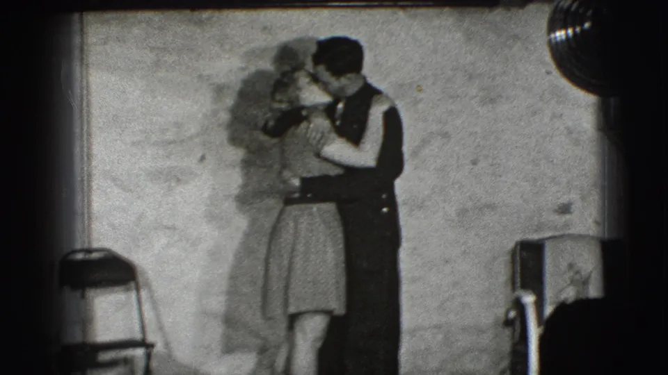 In the supplied 1941 Joliet home-movie clip, a young couple stands close in a dim room, kissing beside a folding chair and plain wall.
