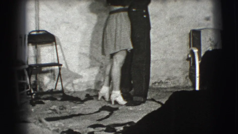 The couple remains close together in the interior, with the woman's light dress and white heels visible near a folding chair and rough floor covering.