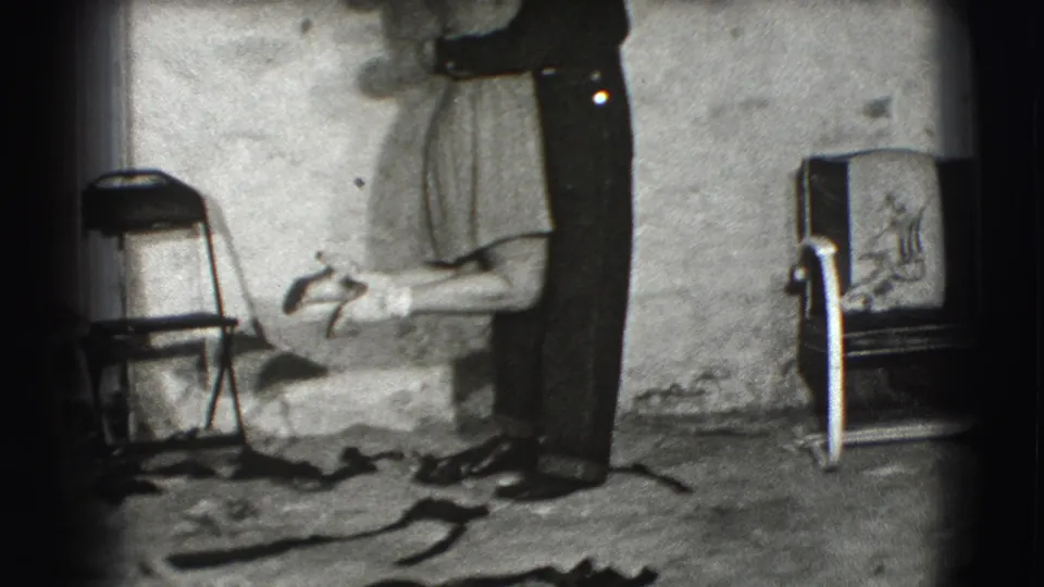 The woman reaches around the man while they stand closely together in the supplied 1941 Joliet interior footage, with a folding chair and wall behind them.