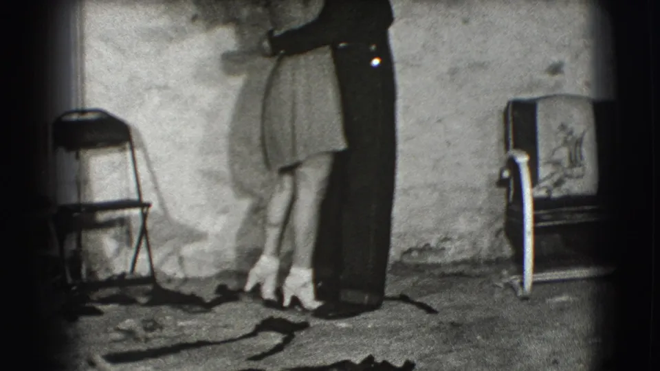 In a 1941 Joliet home-movie frame, a young man embraces a woman in a dim interior with chairs, rough walls, and exposed rafters.