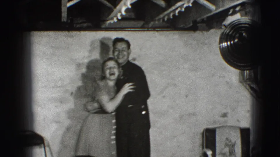 A young man in dark clothing holds a laughing woman in a small Joliet interior, with a chair, wall shadows, and overhead rafters visible.